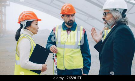 Builders wearing uniform are talking at construction site while architect in suit and helmet is coming discussing project gesturing and looking around Stock Photo