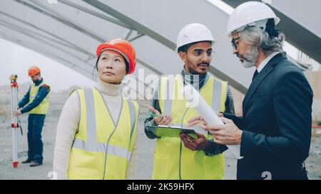 Multiracial group of workers are talking to construction inspector looking at blueprints in building area. Projects and people concept. Stock Photo