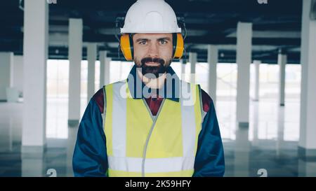 portrait of man builder wearing safety clothing and headphones standing in commercial building smiling looking at camera. Occupational safety and employees concept. Stock Photo