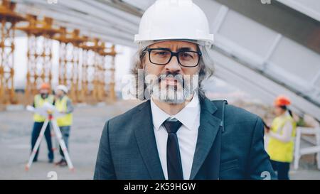 Portrait of serious mature man investor standing outdoors in construction site wearing suit and safety helmet and looking at camera while builders working in background Stock Photo