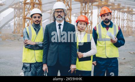 Multi-ethnic group of people builders standing outdoors in industrial site and looking at camera. Team members are wearing uniform. Stock Photo