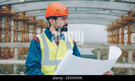 Portrait of serious foreman wearing uniform checking blueprint and looking at construction site outdoors in urban area. People and occupation concept. Stock Photo