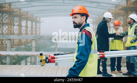 serious male surveyor in uniform walking in construction site holding theodolite concentrated on work. People and profession concept. Stock Photo