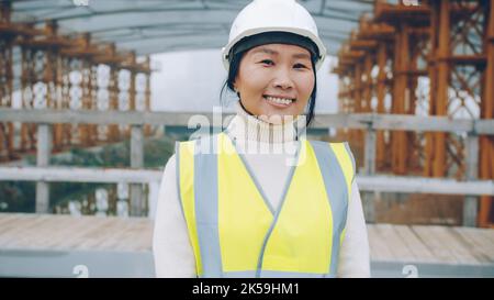 Portrait of cheerful Asial lady wearing helmet and protective vest smiling in building area looking at camera. Women and professions concept. Stock Photo