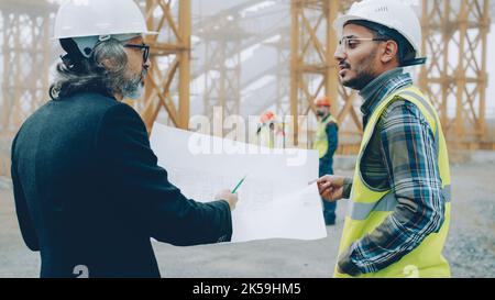 Back view of engineers discussing construction project looking at blueprint while surveyors are busy with equipment in background. Building and management concept. Stock Photo