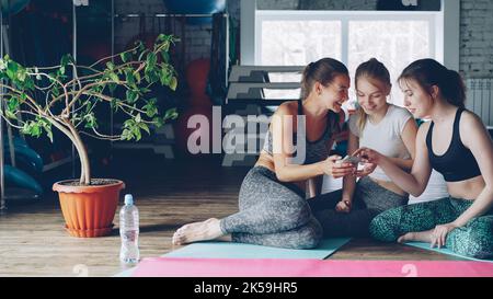 Young attractive women are socialising watching smartphone in gym. They are touching screen, gesturing, chatting emotionally and laughing. Sports and modern technology concept. Stock Photo
