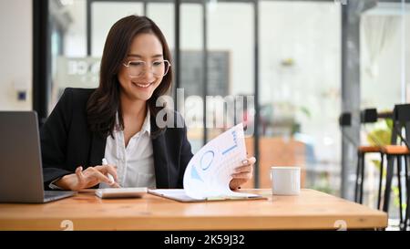 Positive and charming millennial Asian businesswoman or female accountant wearing eyeglasses working at her desk, using calculator to recheck a financ Stock Photo