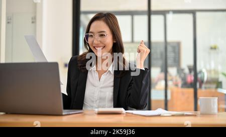Attractive and charming millennial Asian businesswoman or female financial manager in eyeglasses working at her desk, looking at her laptop screen, pl Stock Photo
