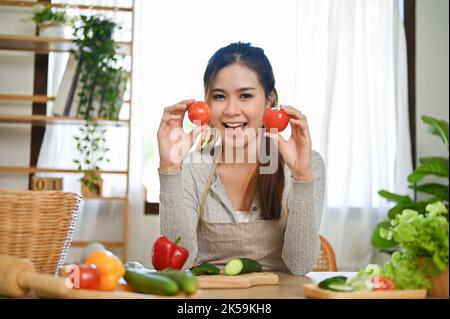 Charming and pretty young Asian female holding tomatoes, smiling and looking at camera, enjoying making a salad bowl in the kitchen.