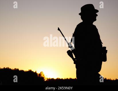 Up at the crack of dawn. A silhouette of a hunter with a rifle on his back and holding binoculars with copy space. Stock Photo