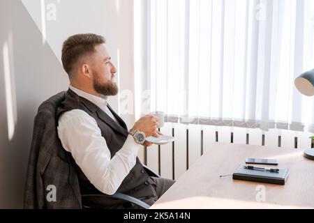 Enjoying fresh coffee at work. Confident young bearded man in business clothes working drinking a hot drink while sitting at his workplace in the office with a pensive look Stock Photo