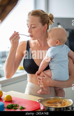 Woman cooking and tasing food while holding four months old baby boy in her hands Stock Photo