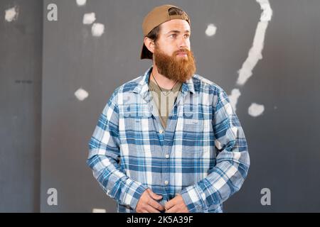 Bearded man in a shirt and a baseball cap on the background of the wall. Repair and renovation home Stock Photo