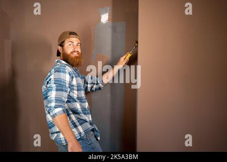 Portrait of happy bearded painter in cap painting a wall with paint roller. House Painter Stock Photo