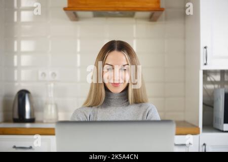 Cheerful blonde woman working on computer in kitchen. Beautiful white femaler doing distant work on laptop at home Stock Photo