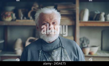 Close-up portrait of senior potter in muddy apron standing alone at table in workshop, smiling and looking at camera. Shelves with handmade vases and pots in background. Stock Photo
