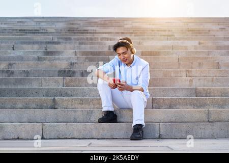 Cheerful young Asian man using mobile phone outdoors, while listening music, sitting on big stairs. Stock Photo