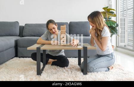 Two asian woman enjoy playing Jenga wooden blocks game in the living room. Players take turns removing one block at a time from a tower constructed of Stock Photo