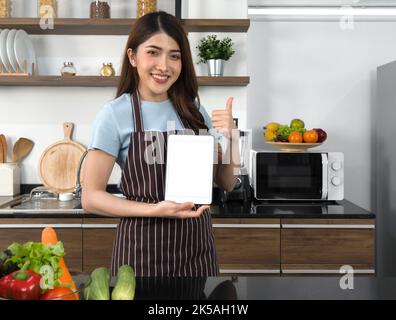 The housewife dressed in an apron  showing white screen tablet computer while raise finger thumbs up. Morning atmosphere in a modern kitchen. Stock Photo