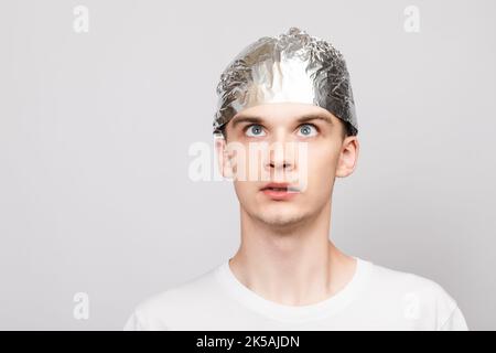 Portrait of anxious young man wearing tin foil hat looking up. Conspiracy theories and paranoya concept. Studio shot on gray background Stock Photo