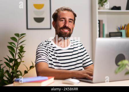 Young caucasian guy looking at camera smiling, sitting at home using laptop to give an online lesson Stock Photo