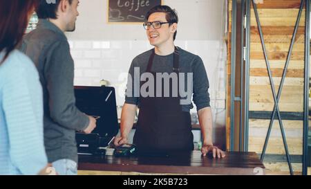 Cheerful friendly cashier is taking orders from customers standing in line, accepting payment and selling takeaway coffee. Stock Photo