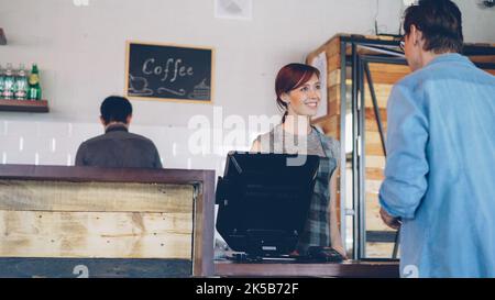 Busy work day at cozy coffeehouse with customer standing at counter buying coffee and friendly employees working and talking. Stock Photo