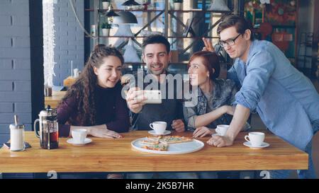 Young bearded man is making video call from pizza house together with best friends. Playful mates are talking, smiling, making funny faces and waving hands looking at camera. Stock Photo