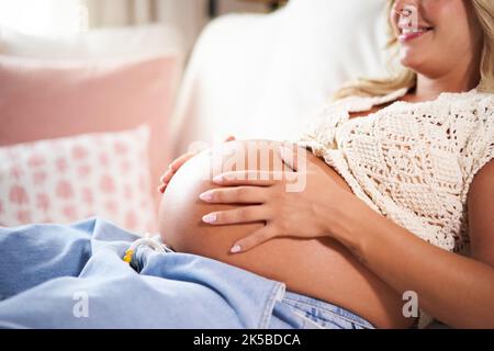 Sweet and happy photo of pregnant lady cuddling her belly while lying on the sofa resting at home. Stock Photo