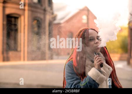 Portrait of young woman with dreadlocks vaping, standing on street. Female with colourful hairstyle smoking e-cigarette, letting off steam Stock Photo
