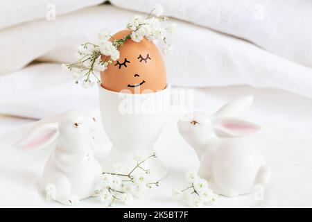 Easter egg with cute face in floral wreath crowns in egg cup with two white easter bunnies. Easter egg with flowers and sleepy eyes in sunny light and Stock Photo