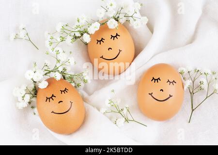 Three happy Easter eggs with cute faces in floral wreath crowns on white background. Easter eggs with flowers and sleepy eyes in sunny light. Happy Ea Stock Photo