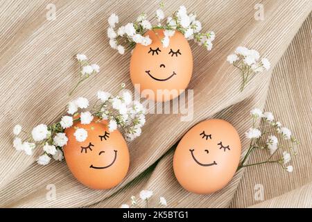 Three happy Easter eggs with cute faces in floral wreath crowns on brown burlap background. Easter eggs with flowers and sleepy eyes in sunny light. H Stock Photo