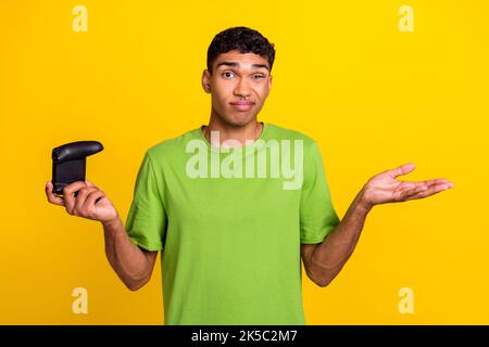 Photo of young handsome funny guy grimace dont know no idea hold wireless joystick player palms problem sorry isolated on yellow color background Stock Photo