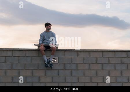 Young man sitting on a wall with a skateboard at sunset Stock Photo
