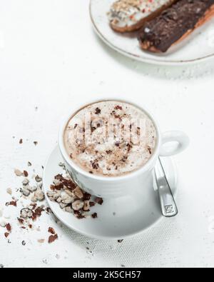 Cappuccino with hazelnuts on a white table and in the corner Traditional French dessert eclair with dark chocolate Stock Photo