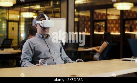 Man using VR headset and wireless controllers, talking online with client, having video call, working in modern office of hi-tech company. Business virtual meeting. Cyberspace digital technology Stock Photo