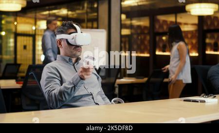 Male office worker wearing VR headset and using wireless controllers, gesturing, watching data in virtual reality. Work in modern office of hi-tech company. Cyberspace digital technology Stock Photo