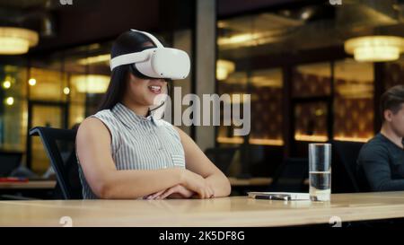 Asian businesswoman wearing VR headset, speaking online with client, having video call, working in modern office of marketing company. Business meeting in virtual reality. Cyberspace and metaverse Stock Photo