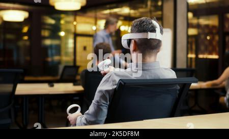 Back view of man wearing VR headset and using wireless controllers, watching data in virtual reality. Work in modern office of hi-tech company with diverse coworkers. Cyberspace digital technology Stock Photo