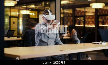 Male office worker wearing VR headset and using wireless controllers, gesturing, watching data in virtual reality. Work in office of hi-tech company near coworkers. Cyberspace digital technology Stock Photo