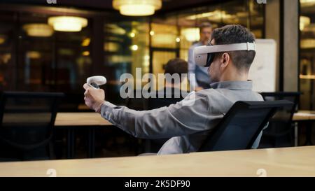 Back view of man wearing VR headset and using wireless controllers, watching data in virtual reality. Work in modern office of hi-tech company with diverse coworkers. Cyberspace digital technology Stock Photo