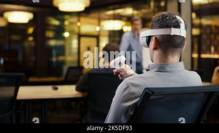 Back view of man wearing VR headset and using wireless controllers, watching data in virtual reality. Work in modern office of hi-tech company with diverse coworkers. Cyberspace digital technology Stock Photo