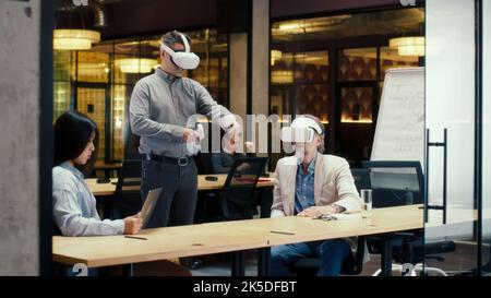 Multi-ethnic office workers testing VR headsets together, using tablet computer and wireless controllers, watching data, working in modern office of hi-tech company. Cyberspace digital technology Stock Photo