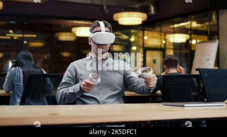 Male office worker wearing VR headset and using wireless controllers, gesturing, watching data in virtual reality. Work in office of hi-tech company near coworkers. Cyberspace digital technology Stock Photo