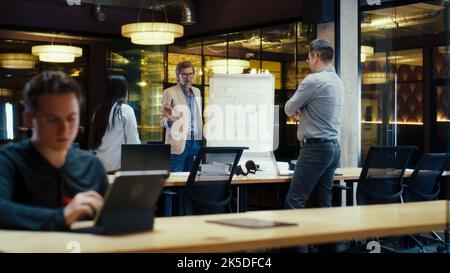 Diverse office workers having meeting, discussing blueprint on flipchart. Presentation of engineering architect startup in modern office of company. Businessman leading conference with managers Stock Photo