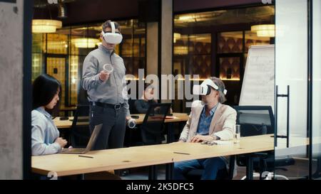 Multi-ethnic office workers testing VR headsets together, using tablet computer and wireless controllers, watching data, working in modern office of hi-tech company. Cyberspace digital technology Stock Photo