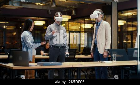 Multi-ethnic office workers testing VR headsets together, using tablet computer and wireless controllers, watching data, working in modern office of hi-tech company. Cyberspace digital technology Stock Photo