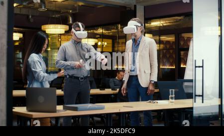 Multi-ethnic office workers testing VR headsets together, using tablet computer and wireless controllers, watching data, working in modern office of hi-tech company. Cyberspace digital technology Stock Photo