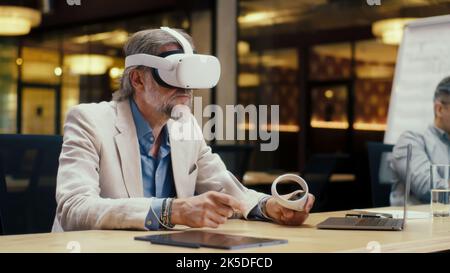 Businessman in VR headset using wireless controllers and laptop, gesturing, watching data in virtual reality. Work in modern office of hi-tech company. Cyberspace digital technology Stock Photo
